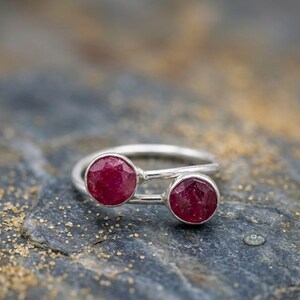 Puede incluir: Un anillo de plata con dos gemas redondas, facetadas, de color rojo intenso. La banda del anillo es un diseño simple y delgado de plata, con las gemas colocadas una al lado de la otra. El anillo se muestra sobre una superficie texturizada de color gris oscuro.