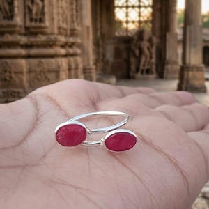 May include: A silver ring featuring two oval-shaped, vibrant red gemstones. The ring's design is open, with the gemstones set on either side of the finger. The ring is displayed on a hand, showcasing the jewelry's design and color.