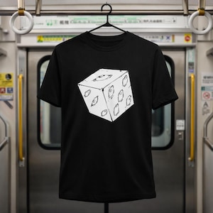 May include: Black t-shirt featuring a white graphic of a cube with multiple eye designs. The cube design is centered on the front of the shirt. The shirt is hanging on a black hanger.
