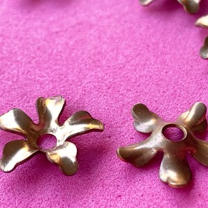 May include: Five gold metal flower-shaped embellishments with a hole in the center, on a pink surface.