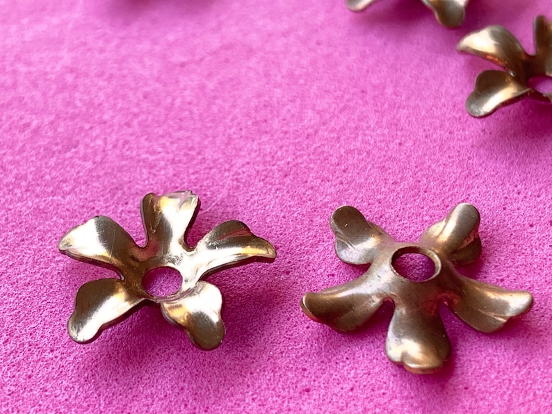 May include: Five gold metal flower-shaped embellishments with a hole in the center, on a pink surface.