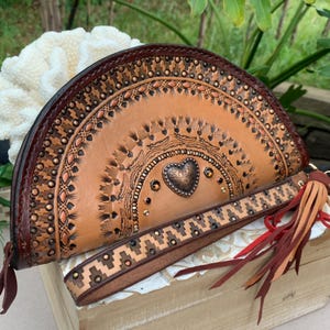 Puede incluir: Un bolso de mano de cuero marrón claro con un diseño en forma de corazón y un borde de cuentas. El bolso tiene una correa larga con un borla.