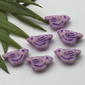 May include: Six lavender bird-shaped beads with purple etched details. The beads have a slightly distressed appearance, with a small eye detail and etched wing and tail features. The beads are arranged on a white surface with green leaves.