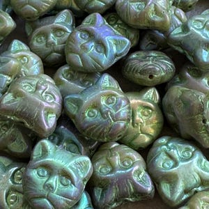 May include: Close-up of iridescent cat-shaped beads in shades of green, blue, and purple. The beads have a detailed cat face design with visible eyes, nose, and whiskers. The beads are clustered together, creating a textured surface. Ideal for jewelry making.