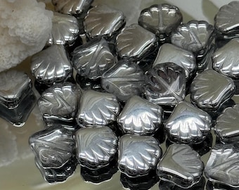 May include: A collection of shiny, silver-colored seashell-shaped beads. The beads have a detailed, textured surface, reflecting light. The image shows a close-up view of the beads, suggesting they are for jewelry making or crafting.