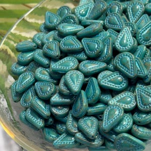 May include: A collection of teal-coloured, teardrop-shaped beads with a textured, embossed border pattern. Each bead features a smooth, slightly glossy finish with subtle variations in colour tone, arranged in a dense, overlapping pile.