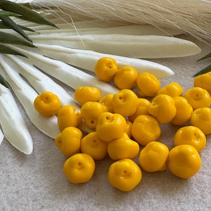 May include: A collection of bright yellow, round beads with a small hole at the top. The beads are scattered on a light gray surface, with white and green decorative elements in the background. These beads are ideal for jewelry making.
