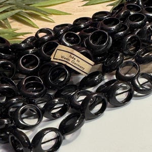 May include: A collection of glossy black circular rings or beads arranged in a cluster. A small rectangular paper label is attached to the center, featuring the printed text Made in Western Germany in black ink on a light tan background.