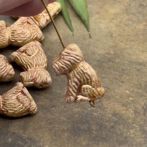 May include: Close-up of several small, tan-coloured dog-shaped beads. The beads have detailed textured surfaces and are strung on a thin wire. The background is a muted, earthy tone.