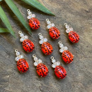 May include: Ten gold-toned ladybug charms with red enamel bodies and clear rhinestone accents. Each charm has a loop for attaching to jewelry or crafts.