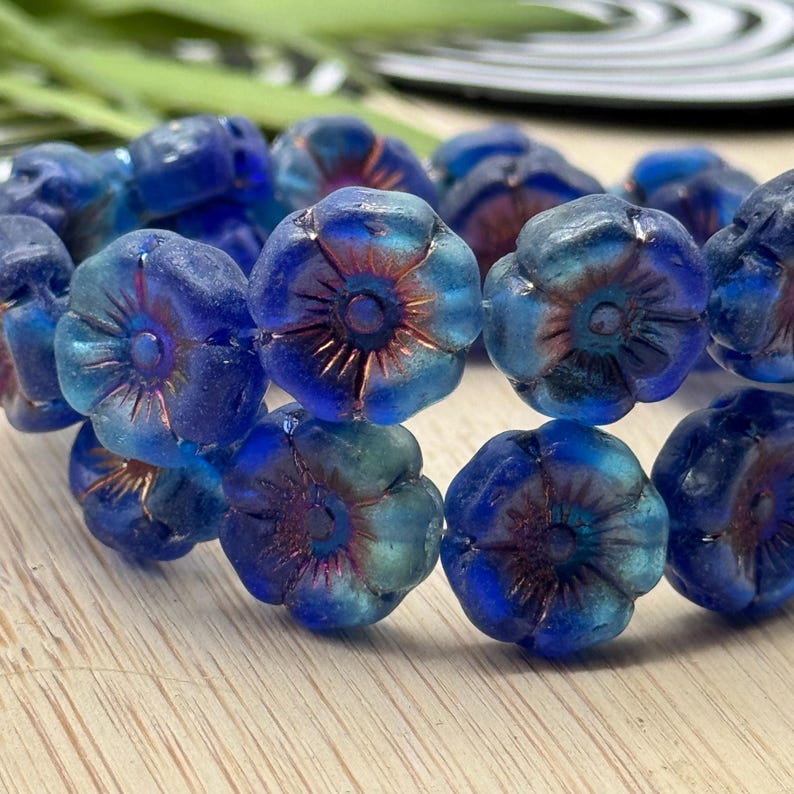 May include: Close-up of a strand of blue flower-shaped beads. The beads have a translucent quality with hints of teal and copper-colored accents in the center. The beads are arranged in a row, suggesting they are part of a bracelet or necklace.