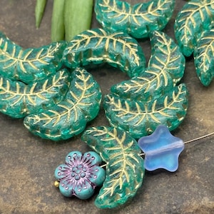 May include: A collection of green glass leaf-shaped beads with gold accents, a blue star-shaped bead, and a turquoise flower bead with purple accents. These beads are ideal for jewelry making and crafting projects.