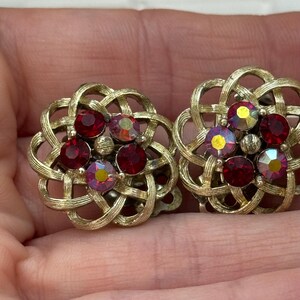 May include: A pair of vintage clip-on earrings. Each earring features a gold-tone, woven metal design with a floral shape. The center of each earring is adorned with red and iridescent rhinestones.