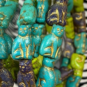 May include: A collection of cat-shaped beads in turquoise, lime green, and dark purple, each featuring gold-coloured accents highlighting the feline features and textured surfaces. The text 2021 WILD DESIGN CO is printed at the bottom right.