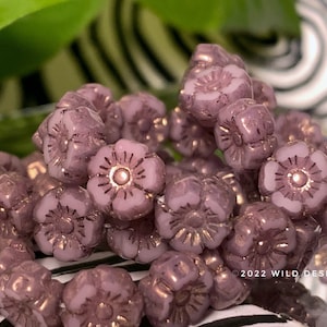 May include: Close-up of a collection of lavender-colored flower-shaped beads. Each bead has a detailed floral design with gold accents. The beads are clustered together, showcasing their intricate details and color variations. The Wild Design Co. copyright is visible.