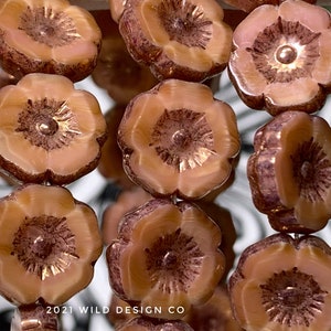 May include: Close-up of several flower-shaped beads. The beads are a light peach color with darker brown accents around the edges and in the center. Each bead has a small, metallic center. The beads are strung together.
