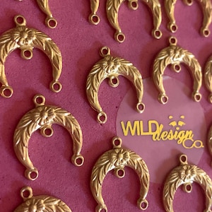 May include: A close-up of multiple gold-toned crescent moon charms with a floral design. Each charm has two loops at the top and two loops at the bottom. The charms are arranged on a pink surface with the text "WILDdesignco." visible in the background.