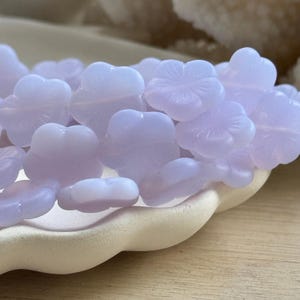 Puede incluir: Un conjunto de cuentas en forma de flor de color lavanda pálido. Las cuentas translúcidas presentan diseños detallados de pétalos. Están dispuestas sobre un plato crema con borde festoneado, lo que sugiere un tema artesanal o de joyería.