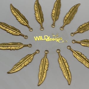May include: Gold-colored feather charms arranged in a circular pattern. Each charm has a detailed feather design with a small loop at the top. The text "WILD Design Co." is visible in the center.