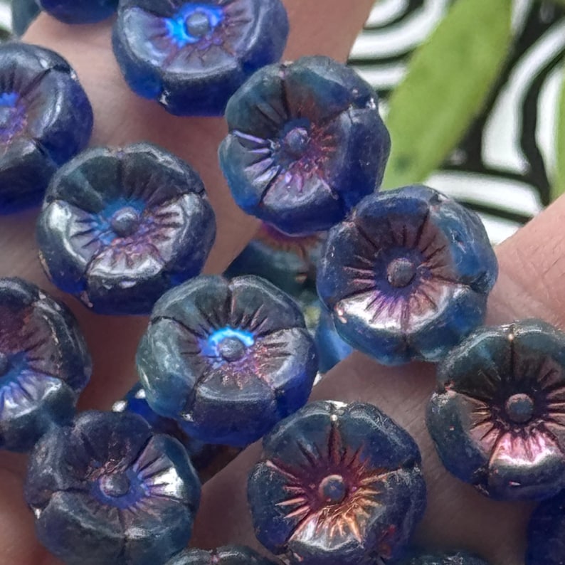 May include: Close-up of several blue flower-shaped beads. Each bead has a detailed petal design and a central, iridescent core. The beads have a slightly translucent quality, with hints of purple and blue hues. These beads are ideal for jewelry making.