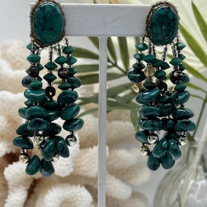 May include: A pair of vintage dangle earrings. Each earring features a large oval, teal-colored stone set in a gold-toned frame. Below, cascading strands of teal beads, black beads, and silver-toned accents create a layered, eye-catching design. The earrings are displayed on a white stand.