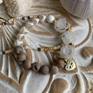 Könnte beinhalten: Ein weißes und braunes Perlenarmband mit einem goldenen Herzanhänger. Das Armband besteht aus weißen Achatperlen, braunen Lavasteinen und klaren, blumenförmigen Perlen.