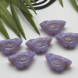 May include: Six lavender bird-shaped beads with etched details. The beads are arranged on a white surface, with green leaves in the background. The bird beads have a small hole for stringing.