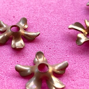 May include: Four gold-colored metal flower-shaped embellishments with a hole in the center. The embellishments are on a pink surface.