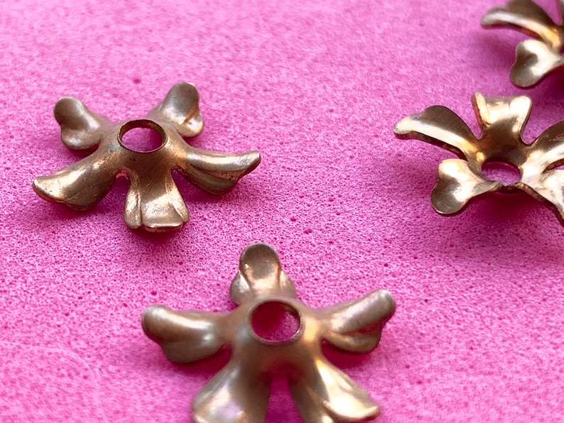 May include: Four gold-colored metal flower-shaped embellishments with a hole in the center. The embellishments are on a pink surface.