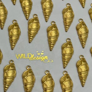 May include: Close-up of multiple gold-colored seashell charms with a loop at the top for attaching to jewelry. The charms are arranged on a white surface. The text "WILD Design Co." is visible in gold.
