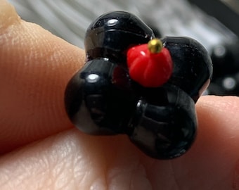 Puede incluir: Una cuenta negra en forma de flor con un centro rojo y un acento dorado. La cuenta está hecha de arcilla polimérica y tiene un diámetro de aproximadamente 2,5 cm.