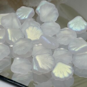 May include: A collection of iridescent, seashell-shaped beads. The beads are a translucent white with a subtle, shimmering effect, catching the light to display hints of blue and yellow. These beads are ideal for jewelry making.