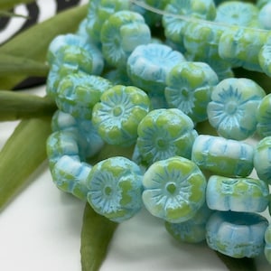 May include: Close-up of a strand of light blue and green flower-shaped beads. The beads have a matte finish and are strung together. The beads are approximately 0.5 inches in diameter. The background is blurred, with green leaves visible.