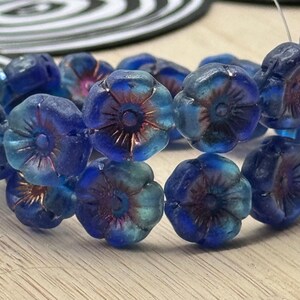 May include: Close-up of a strand of blue flower-shaped beads. The beads have a gradient of colors, from deep blue to turquoise, with a reddish-brown center. The beads are strung together, suggesting they are for jewelry making.