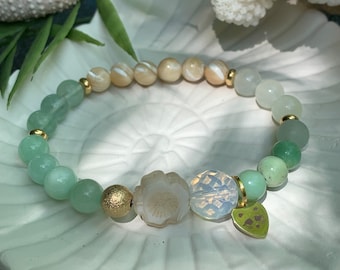 May include: A green and white beaded bracelet with a gold charm and a flower-shaped bead. The bracelet is on a white surface.