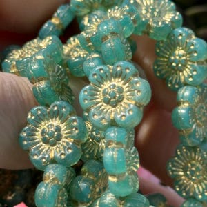 May include: A strand of teal blue glass beads with gold accents. Each bead is shaped like a flower with a raised center and a textured surface.