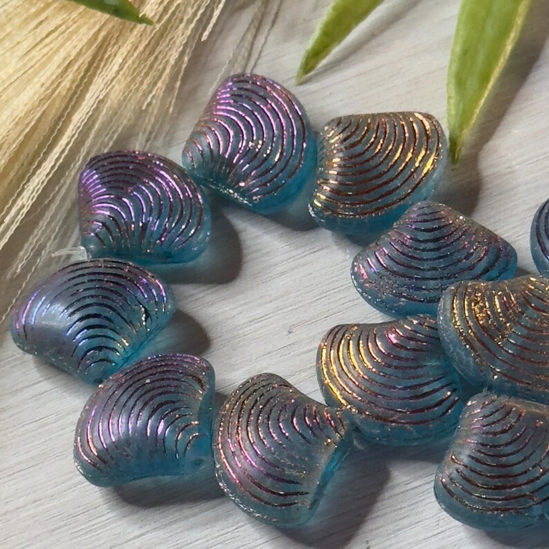 May include: A collection of iridescent blue glass beads shaped like seashells with a gold and purple shimmer. The beads have a textured surface and are ideal for jewellery making.