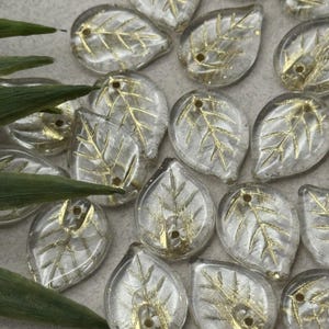 May include: Clear glass leaf-shaped beads with gold leaf vein detailing. Each bead has a small hole for stringing. The beads are scattered on a light gray surface, with green leaves in the background. Ideal for jewelry making.