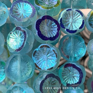 May include: Close-up of a strand of turquoise blue glass beads with a flower design and iridescent finish. The beads have a raised, textured center and a purple rim.