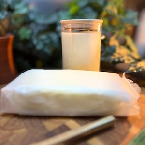 May include: A glass jar with a wooden lid filled with a creamy liquid, next to a rectangular block of wrapped white product. The items are on a wooden surface, with greenery in the background.