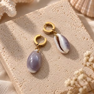 Könnte beinhalten: Ein Paar goldfarbene Creolen, jeweils mit einem hängenden, glatten, ovalen Muschel-Anhänger. Die Muscheln weisen einen sanften, polierten Farbverlauf in Lavendel und Weiß auf und sind an kreisförmigen Metallringen mit sicherem Verschluss befestigt.