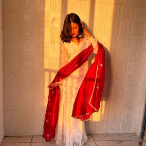 May include: A white ensemble with a red scarf. The outfit includes a long-sleeved top and a matching skirt or pants. The red scarf has a gold trim and is draped over the outfit. The background is a textured wall.
