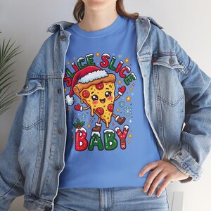 Könnte beinhalten: Ein hellblaues T-Shirt mit einer Grafik eines lächelnden Pizzastücks, das eine Weihnachtsmütze und Stiefel trägt. Der Text SLICE SLICE BABY steht in roten und grünen Buchstaben, umgeben von Schneeflocken, Sternen und Stechpalmen.