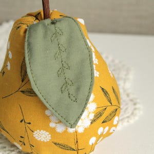May include: A yellow fabric pear with a green leaf design. The pear is decorated with white floral embroidery.