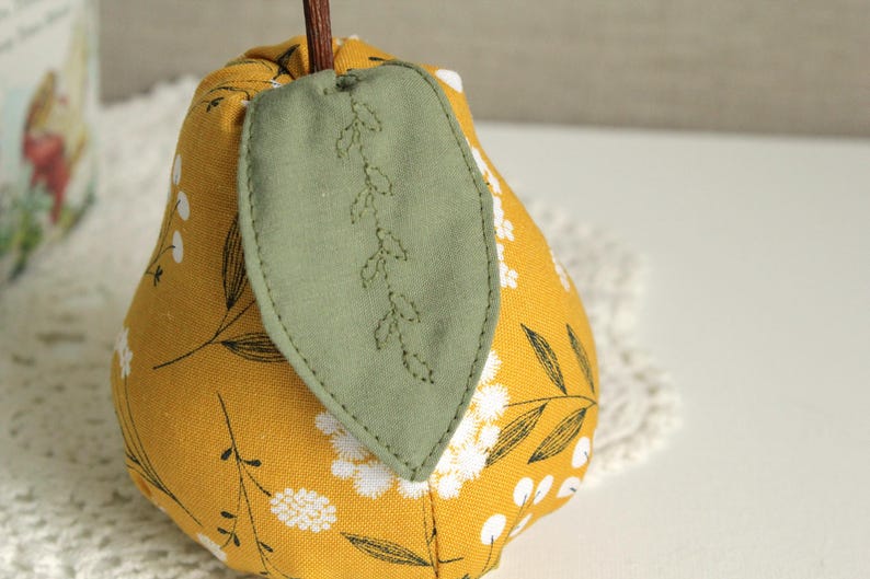 May include: A yellow fabric pear with a green leaf design. The pear is decorated with white floral embroidery.