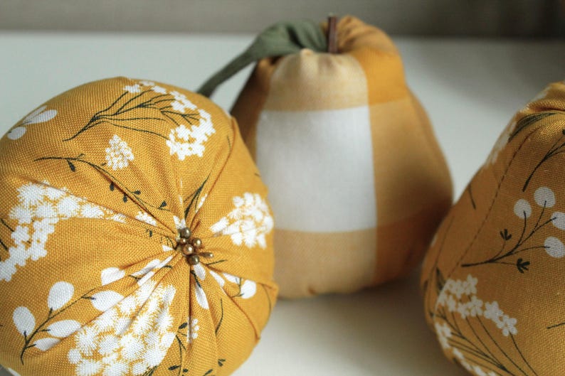May include: Three decorative fabric pumpkins in a yellow and white floral pattern. The pumpkins are made of a soft, textured fabric and have a slightly rounded shape.
