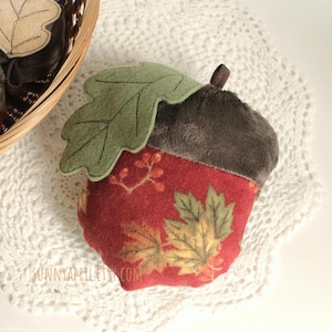 May include: An acorn-shaped decorative item with a plush, dark gray top and a red base adorned with fall leaves and berries. A green felt leaf is attached. The item is on a white lace doily, with similar items in a wicker basket.
