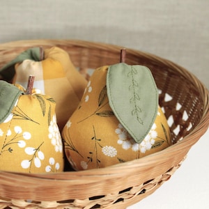 May include: Three fabric pears in a wicker basket. The pears are yellow with different patterns, including a plaid pattern and a floral pattern. Each pear has a green leaf sewn on top.