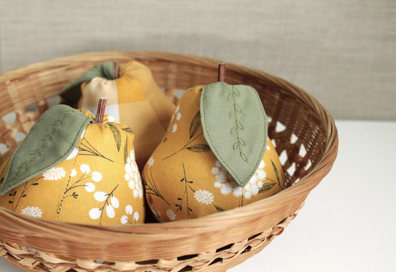 May include: Three fabric pears in a wicker basket. The pears are yellow with different patterns, including a plaid pattern and a floral pattern. Each pear has a green leaf sewn on top.