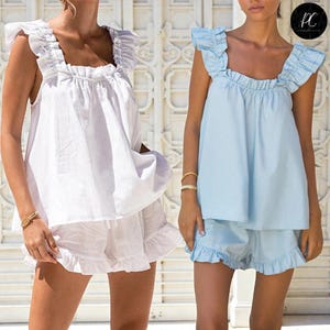 Può includere: Due completi coordinati di pigiami con finiture a balze. Uno è bianco, l'altro azzurro. Ogni completo include un top con scollo quadrato con maniche a balze e pantaloncini abbinati. Gli outfit sono realizzati in tessuto leggero.